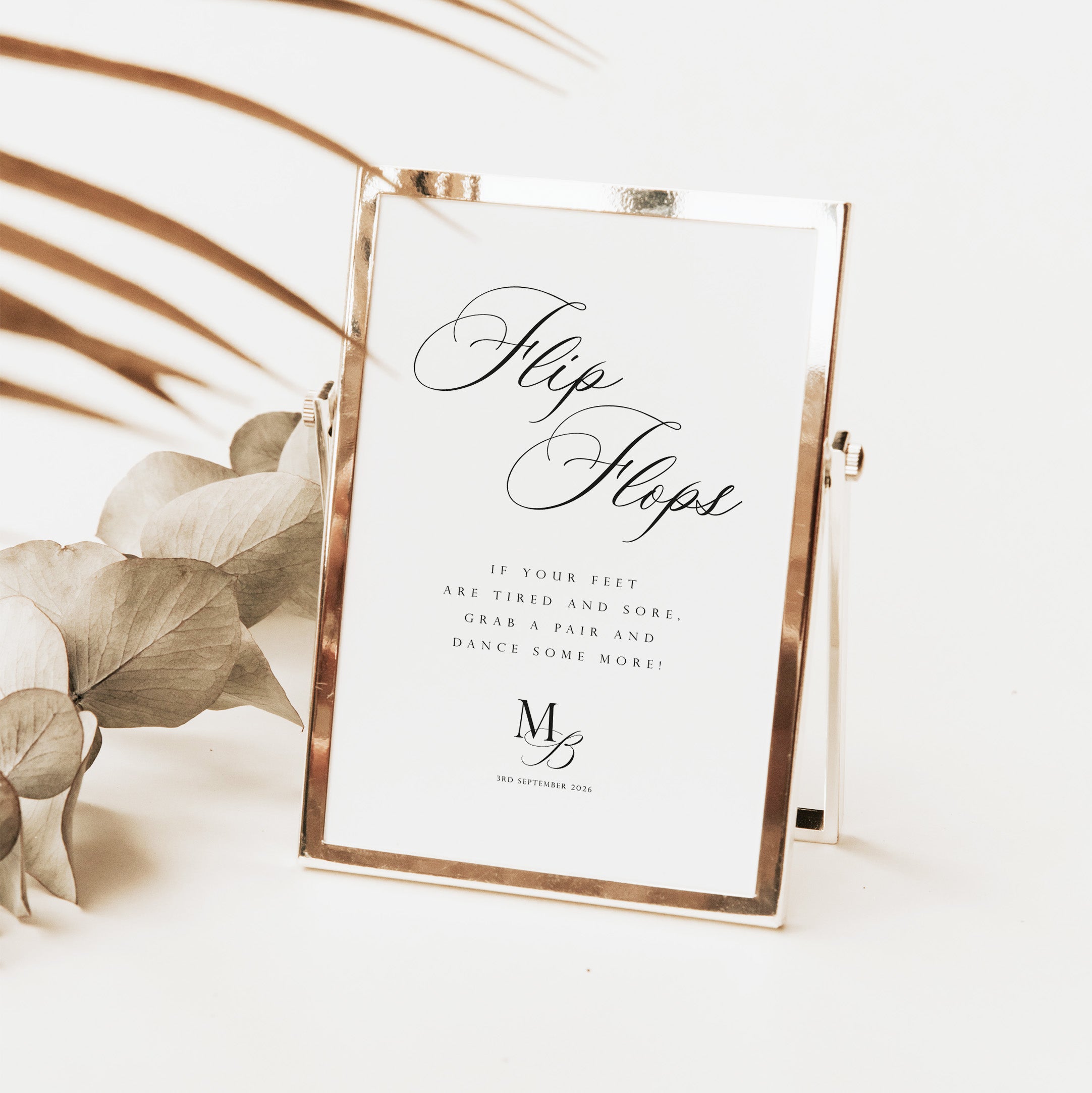 A rectangular tabletop mirror with a thin metallic frame holding a white sign that reads 'FlipFlops' in elegant script, styled on a light surface with dried leaves and palm fronds in the background.