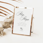 A rectangular tabletop mirror with a thin metallic frame holding a white sign that reads 'FlipFlops' in elegant script, styled on a light surface with dried leaves and palm fronds in the background.
