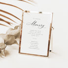 A rose gold picture frame holding a white wedding guest book with elegant black script and serif text, styled on a white surface with dried palm leaves and eucalyptus branches.