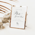 A rose gold metal picture frame standing upright on a white surface, holding a white wedding guest book insert with elegant black script text. Decorative dried leaves and palm fronds are arranged around it in a bright, clean setting.