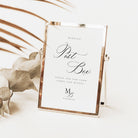 A rectangular wedding post box sign with a metallic rose gold frame and a white insert, styled on a light surface with dried botanical leaves in the background.