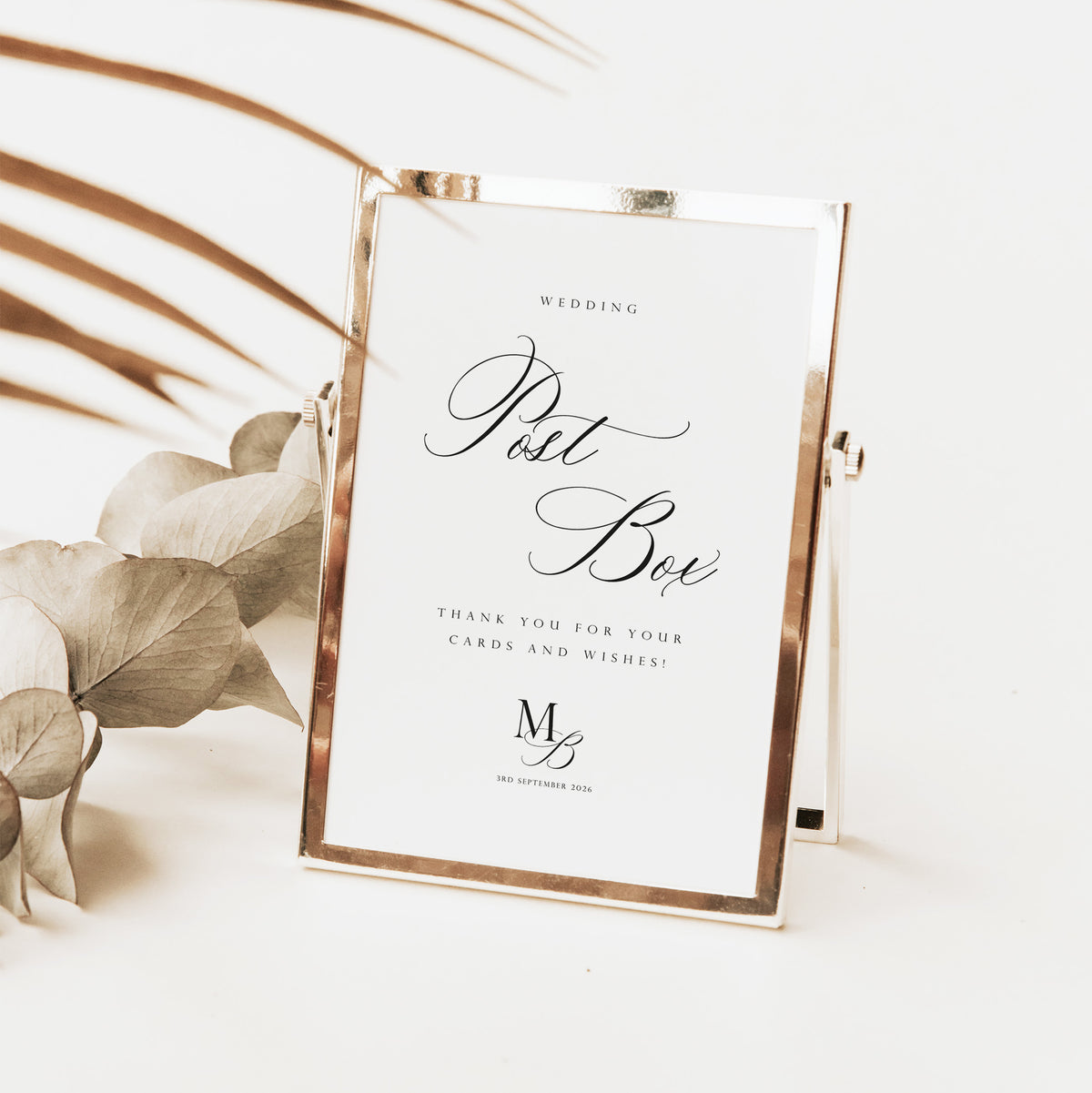 A rectangular wedding post box sign with a metallic rose gold frame and a white insert, styled on a light surface with dried botanical leaves in the background.