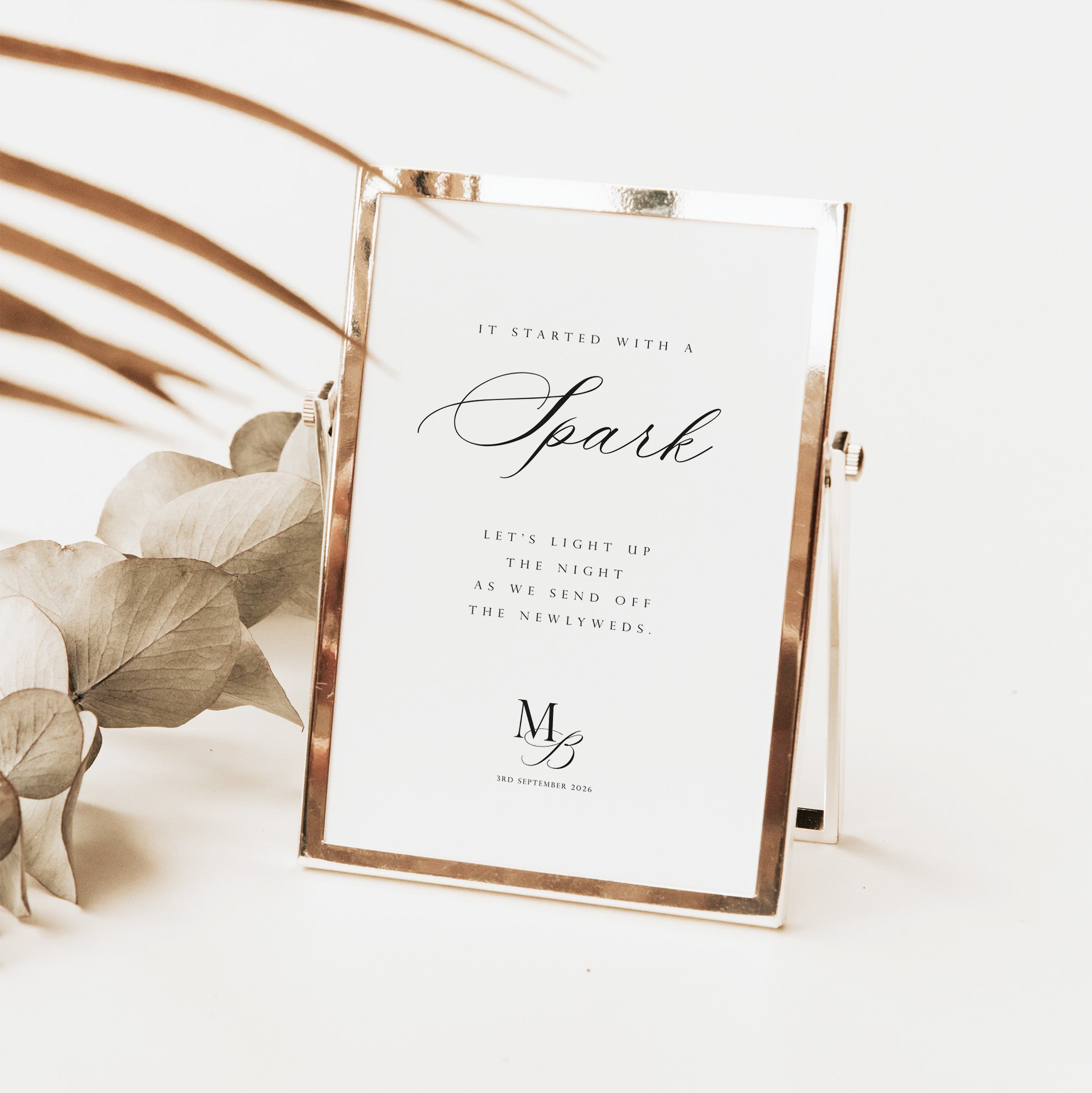 A rectangular photo frame with a metallic rose gold border holding a white printed card for wedding sparklers with elegant black text, styled on a light surface with dried botanical leaves in the background.