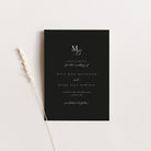 A minimalist black wedding save the date card with elegant white script text, styled flat-lay on a light beige background with a dried wheat stalk.