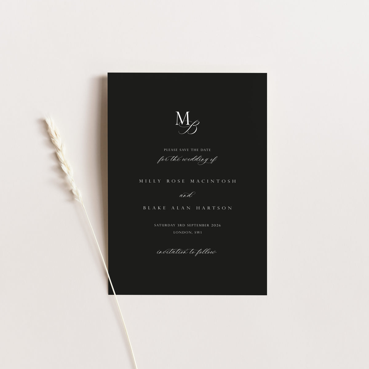 A minimalist black wedding save the date card with elegant white script text, styled flat-lay on a light beige background with a dried wheat stalk.