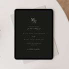 A black tablet with elegant white wedding invitation text and monogram lettering, styled on top of white envelopes on a light neutral surface with a beige fabric corner visible.