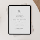 A black tablet displaying a minimalist wedding save-the-date card with elegant black typography on a white page, styled on top of a light gray envelope and a beige textured surface.