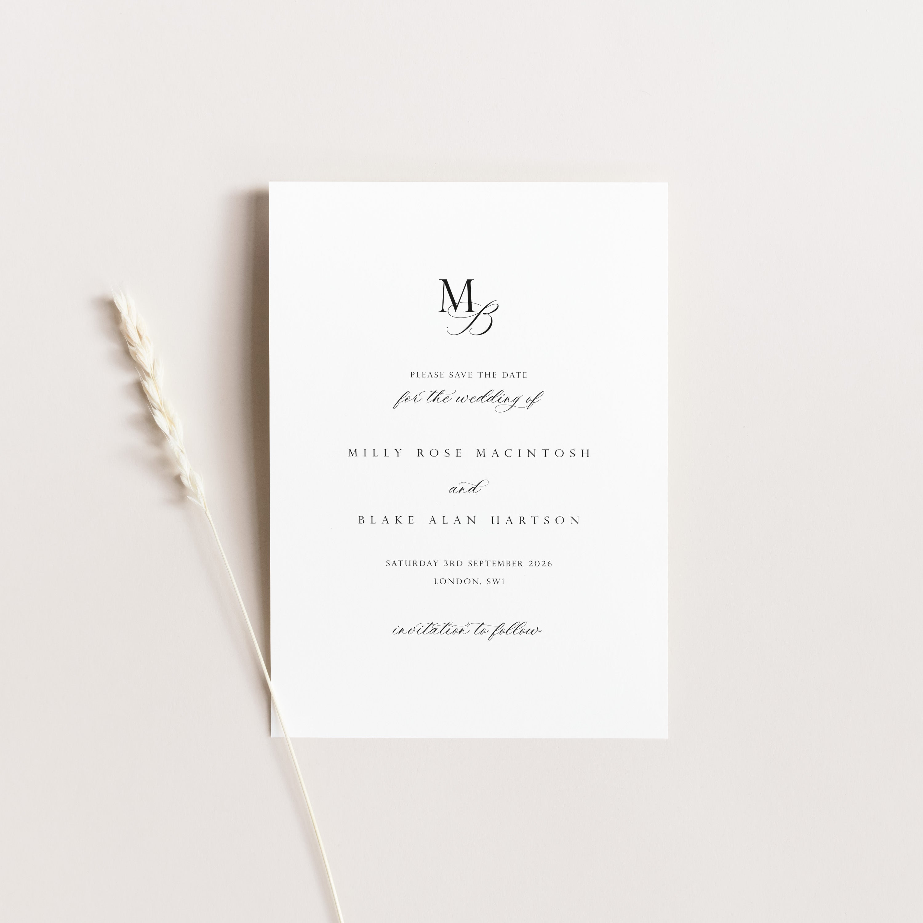 A minimalist wedding save the date card with elegant black typography on a clean white background, styled flat-lay with a dried wheat stem on a warm off-white surface.
