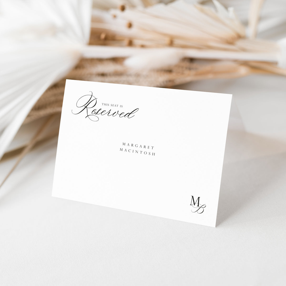 A white reserved seating card with elegant black script text reading 'This seat is reserved' and the names 'Margaret Macintosh,' photographed standing upright on a light surface with soft dried floral decor in the background.