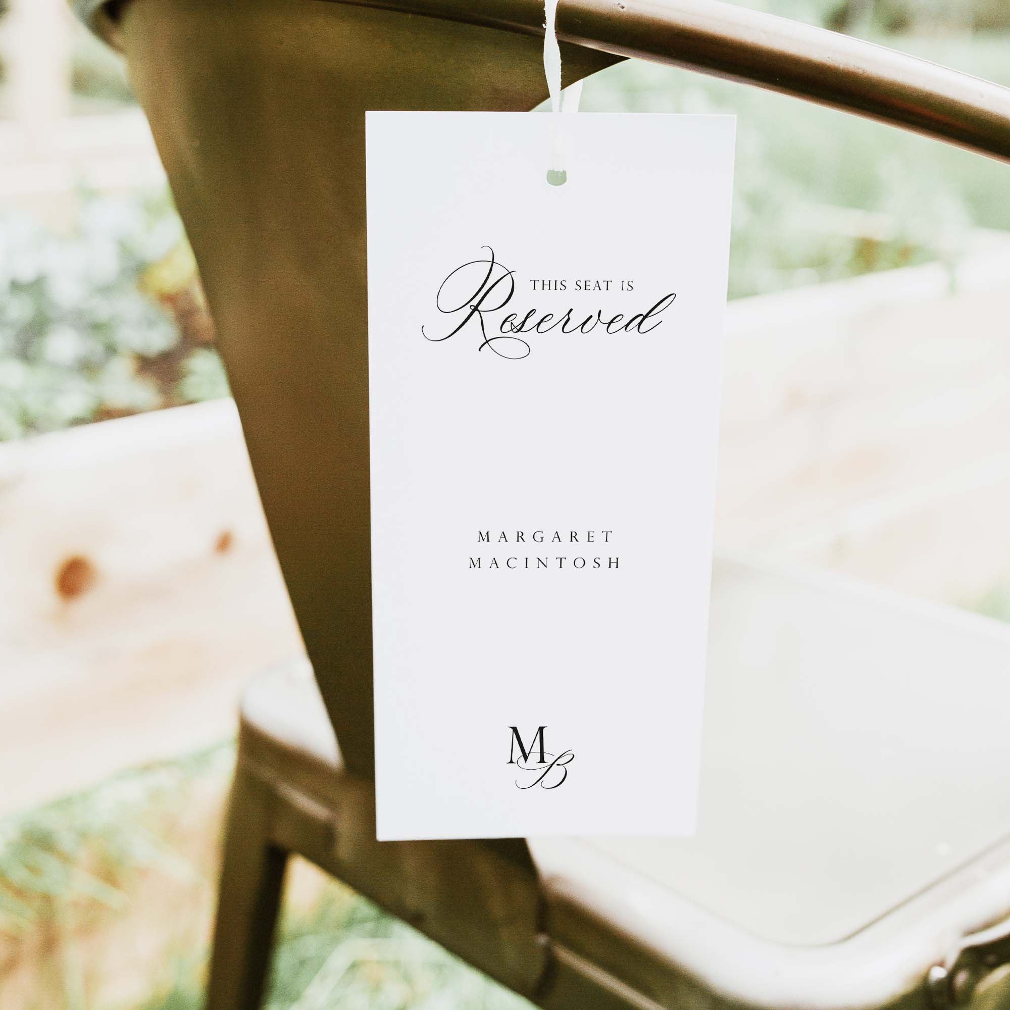 A white reserved seat tag hanging from a metal chair in an outdoor setting. The tag features elegant black script reading 'THIS SEAT IS Reserved' and the names 'MARGARET MACINTOSH' with a monogram logo at the bottom.