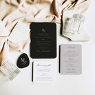 A styled flat-lay of a wedding invitation suite on a white surface with draped blush fabric. The set includes a black invitation card, a white 'The morning after' card, a light grey details card, a black tag with an MB monogram, and clear ribbed glasses.