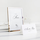 A rose gold metal table number frame standing upright next to a white paper table card on a clean white surface with soft decorative branches in the background.