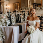 Woman in a white wedding dress sitting in a decorated room with floral arrangements and a chandelier.