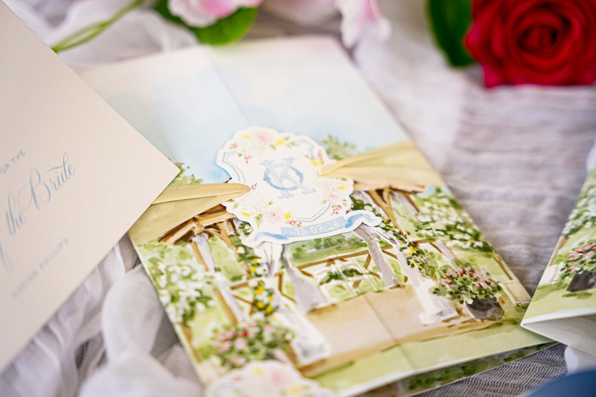 A close-up of a gatefold wedding invitation featuring a watercolour illustrated garden arch scene on neutral fabric and soft pink and red roses in the background.
