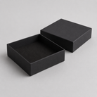 A pair of matte black square gift boxes, one open to show the lined interior and one closed, set against a neutral light gray background.