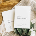 Two white save the date cards displayed on white fabric and a wooden board alongside green foliage.