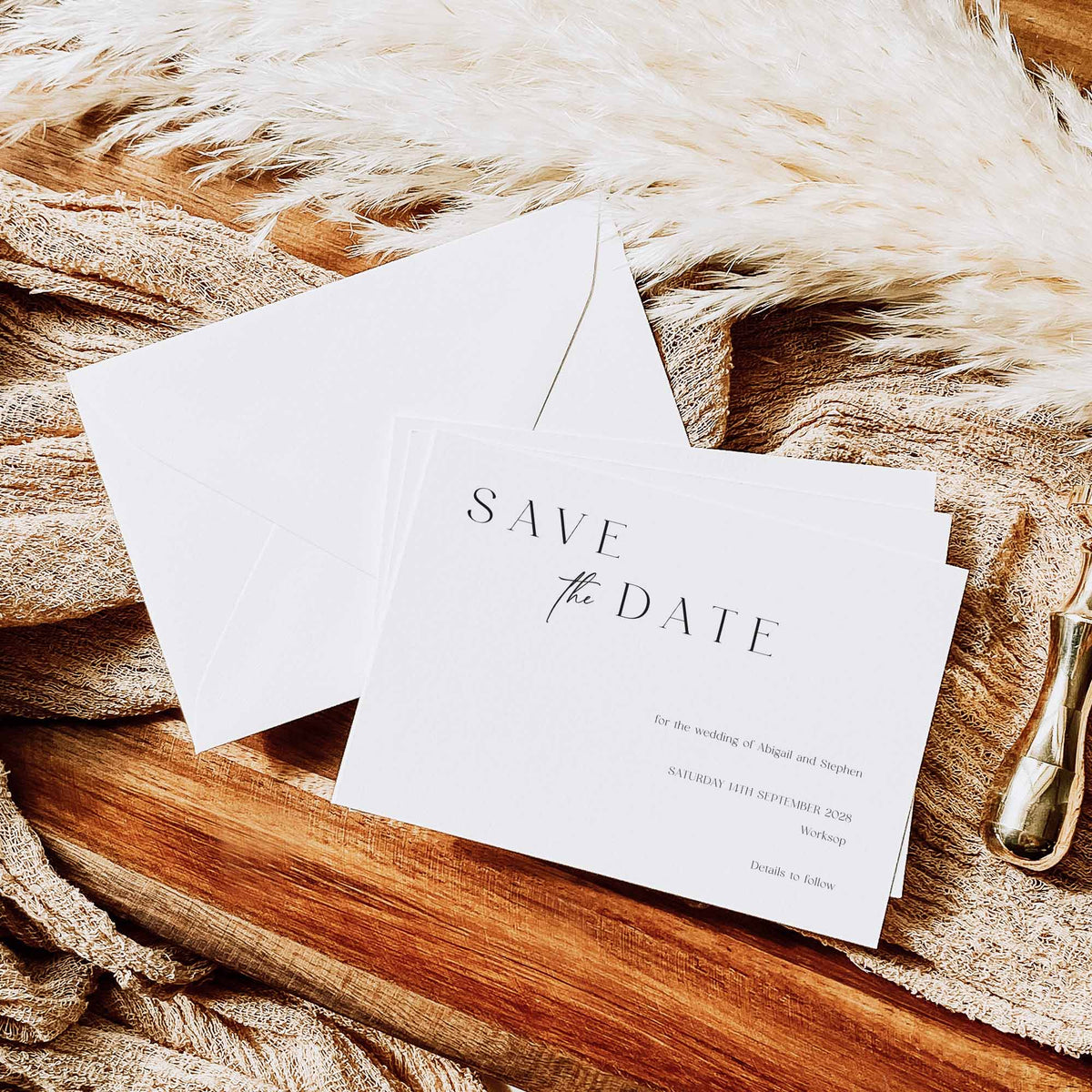 A bundle of white save the date cards shown above a white diamond flap envelope on a wooden board alongside neutral fabric, wax stamp and pampass grass.