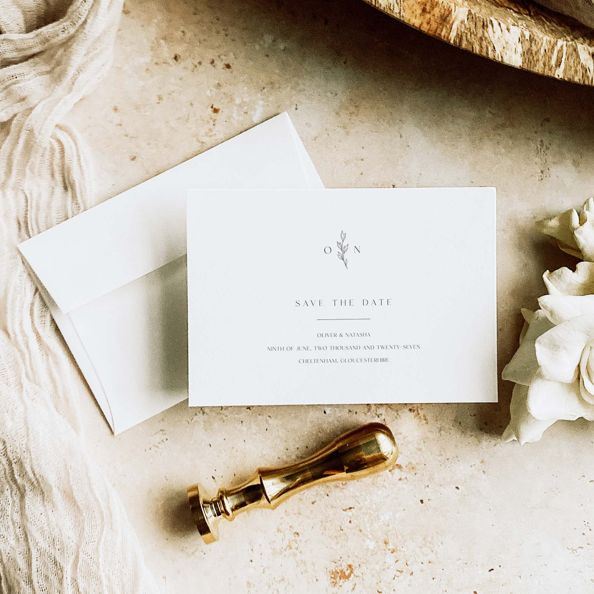 A styled flat-lay of a white 'Save the Date' card with matching white envelope, a gold wax seal stamp, and cream flowers on a textured beige surface.