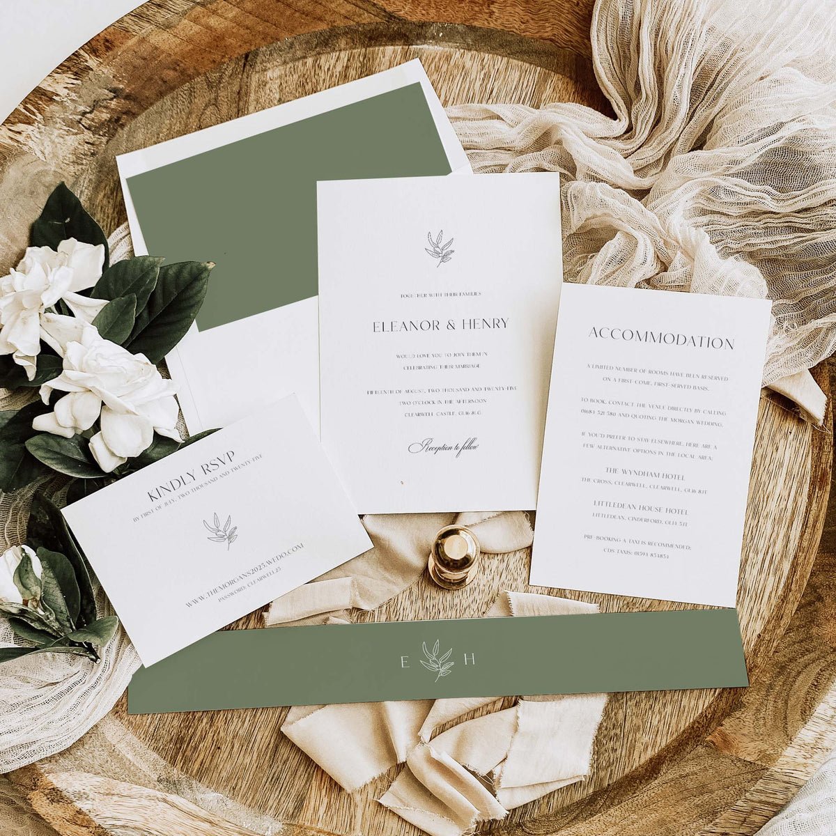 A styled flat-lay of a wedding invitation suite on a rustic wood slice background, including a main invitation card, accommodation card, RSVP card, envelope, green paper bands, cream ribbon, white flowers, and a small gold ring box.