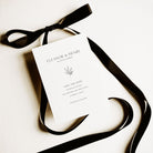 A white save the date card shown on a white coloured background alongside black ribbon tied into a bow