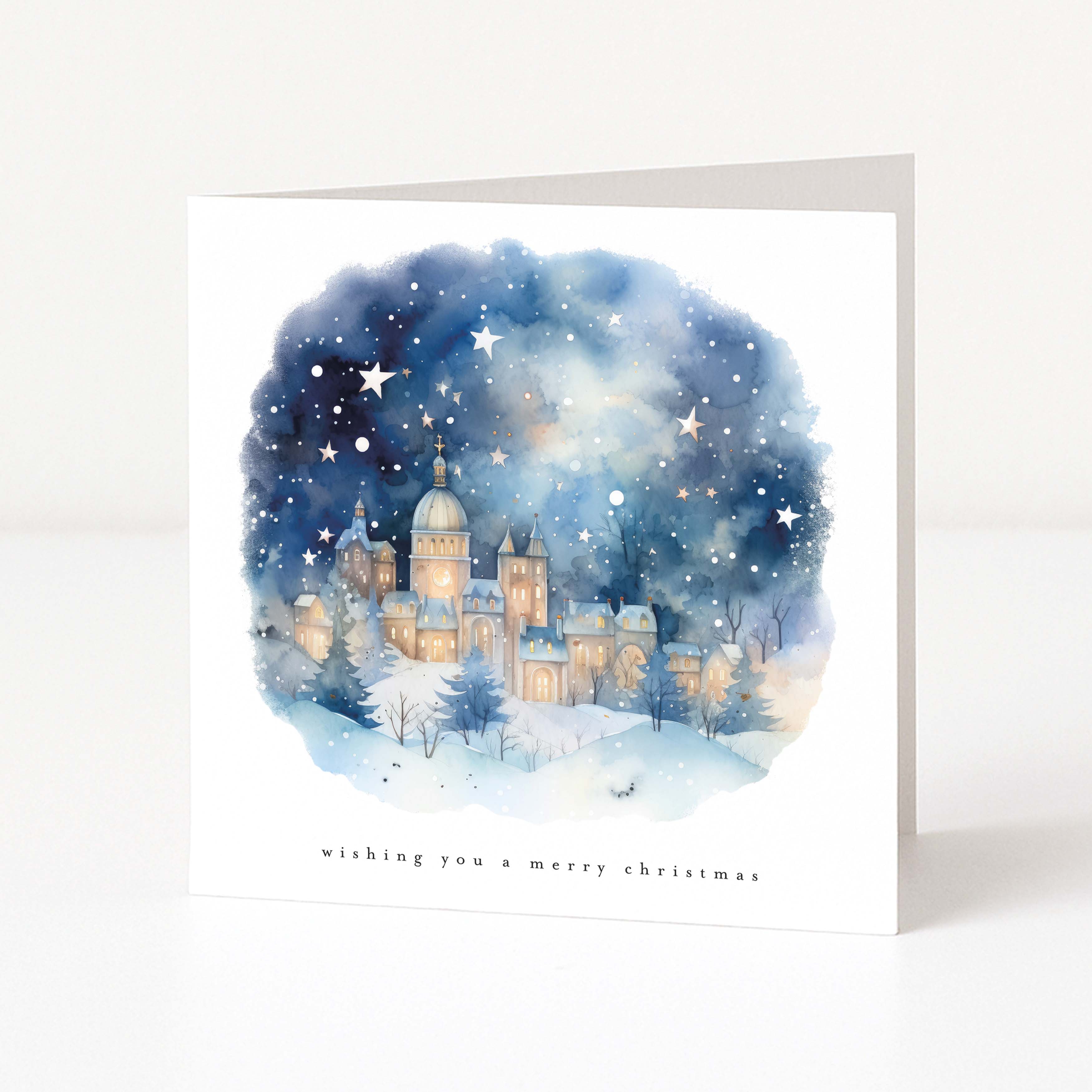 A square white Christmas greeting card standing upright on a white surface. The front features a watercolour illustration of a snowy castle at night under a starry blue sky, with the text 'wishing you a merry christmas' printed below.