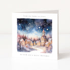 A white Christmas greeting card standing upright on a light gray surface. The front features a watercolor illustration of a snowy village at night under a starry sky, with the text 'from our home to yours' at the top and 'we wish you a merry christmas' at the bottom.