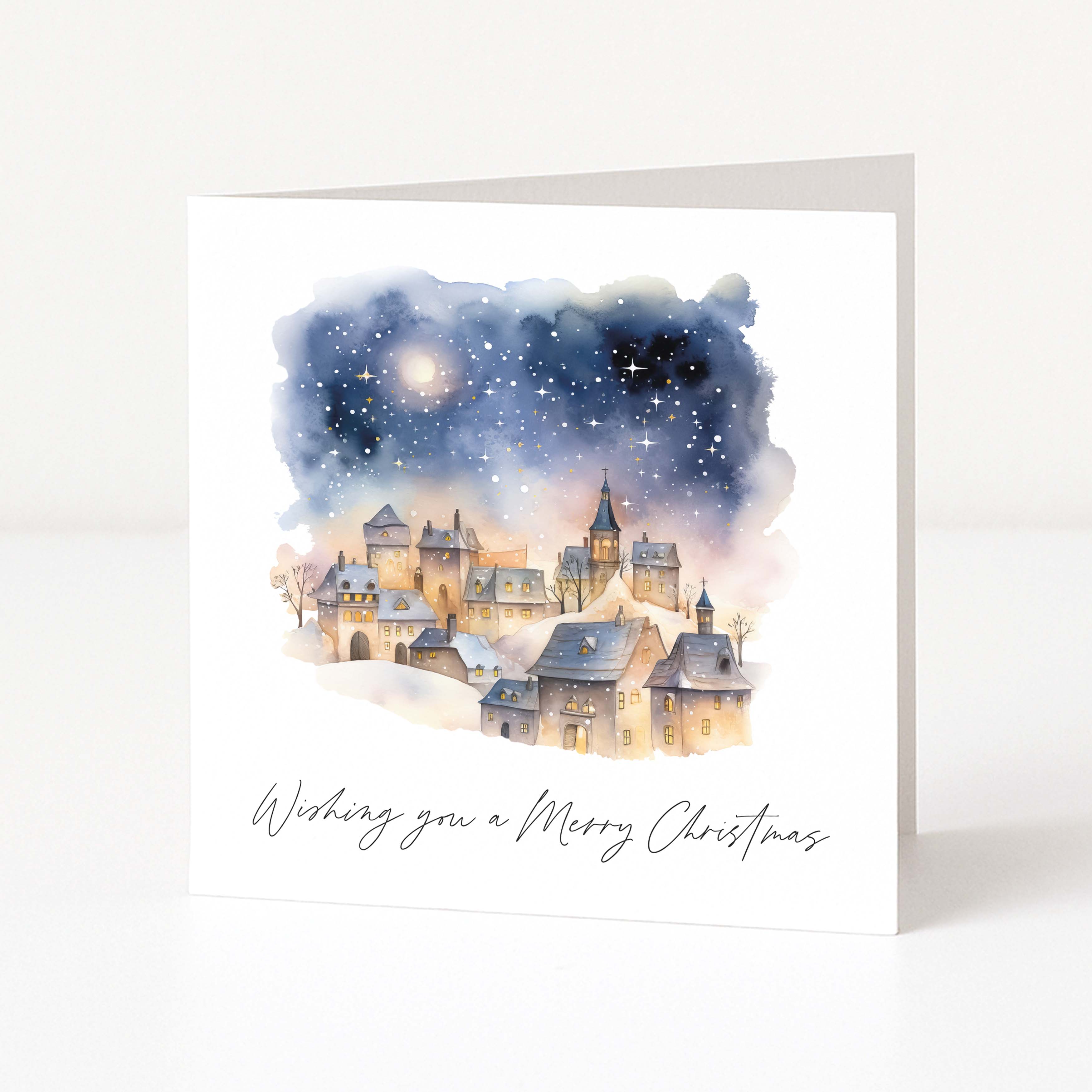 A square white greeting card standing upright, featuring a watercolor illustration of a snowy village under a starry night sky, with the text 'Wishing you a Merry Christmas' printed below.