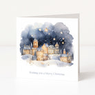 A square white greeting card standing upright on a light surface, printed with a watercolor winter village scene under a starry night sky and the text 'Wishing you a Merry Christmas'.