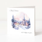 A square Christmas greeting card standing upright on a white surface. The card features a watercolor winter village scene with snow-covered rooftops, lit windows, bare trees, and a soft pink and blue sky.