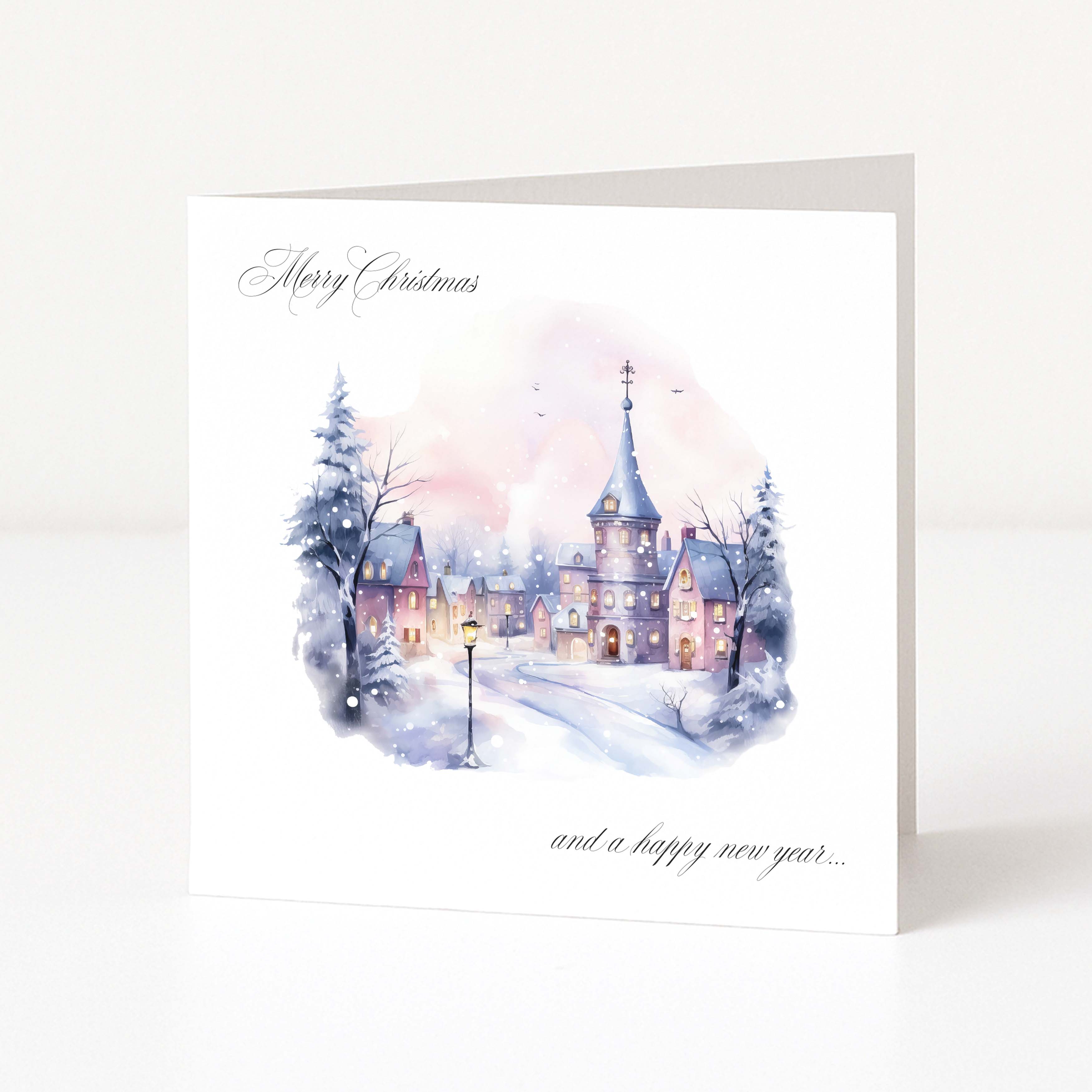 A square Christmas greeting card standing upright on a white surface. The card features a watercolor winter village scene with snow-covered rooftops, lit windows, bare trees, and a soft pink and blue sky.