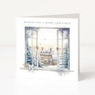 A white Christmas greeting card standing upright, printed with the text 'WISHING YOU A MERRY CHRISTMAS' above an illustrated winter village scene seen through open windows.