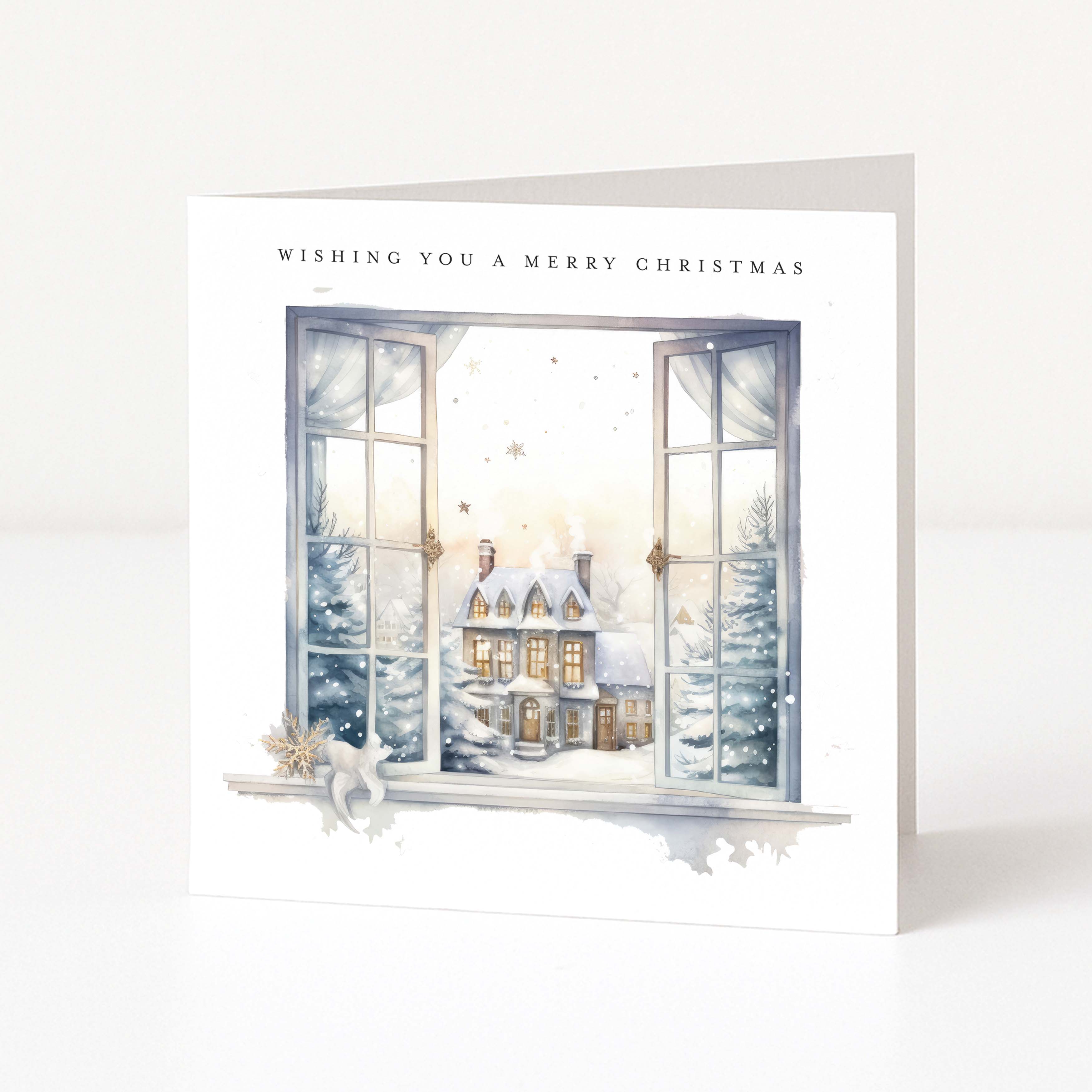 A white Christmas greeting card standing upright, printed with the text 'WISHING YOU A MERRY CHRISTMAS' above an illustrated winter village scene seen through open windows.