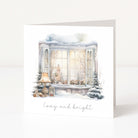 A square greeting card standing upright on a white surface, featuring a watercolor illustration of a snowy bay window with warm glowing windows, a lamp, small trees, and the words 'Cozy and bright' in script below.