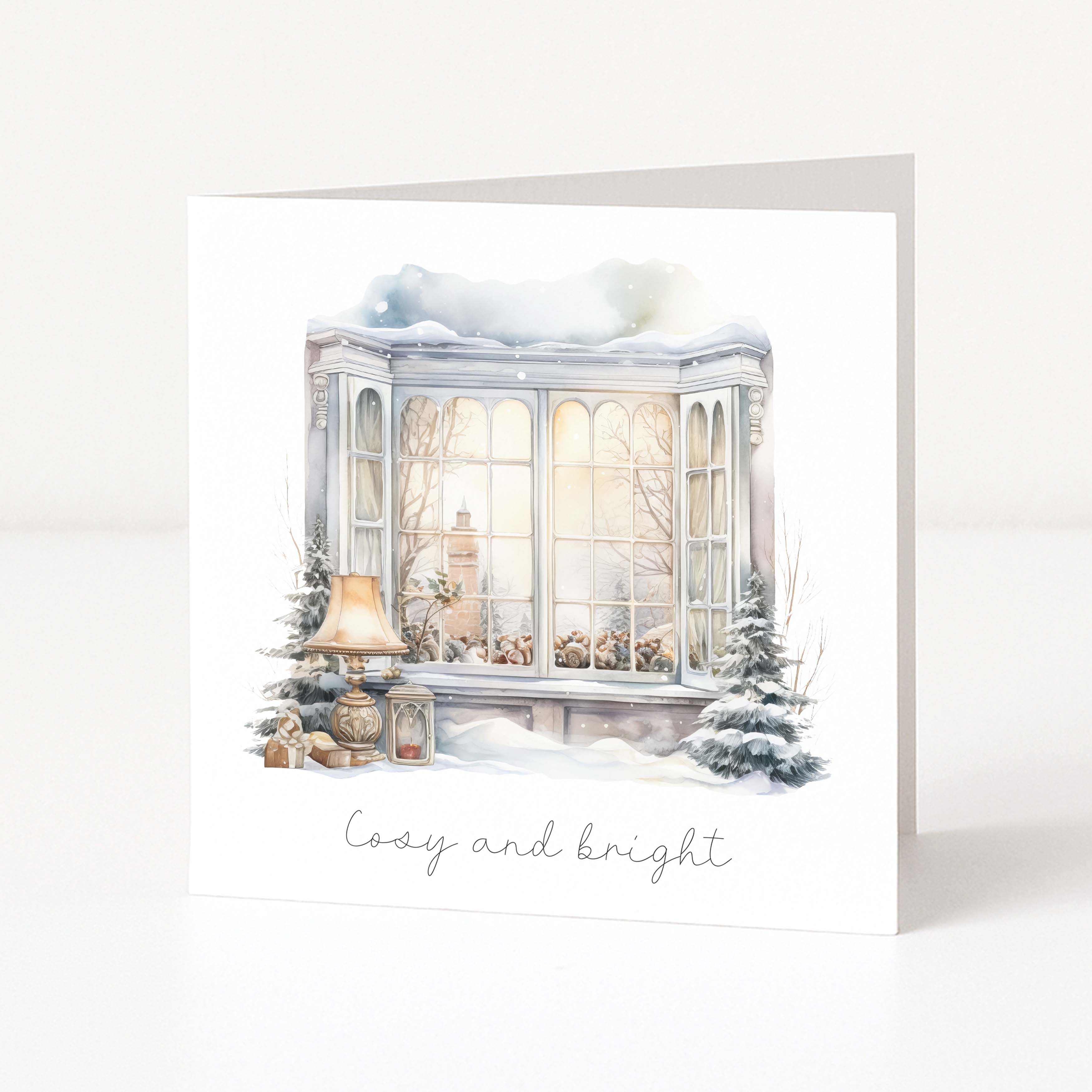 A square greeting card standing upright on a white surface, featuring a watercolor illustration of a snowy bay window with warm glowing windows, a lamp, small trees, and the words 'Cozy and bright' in script below.