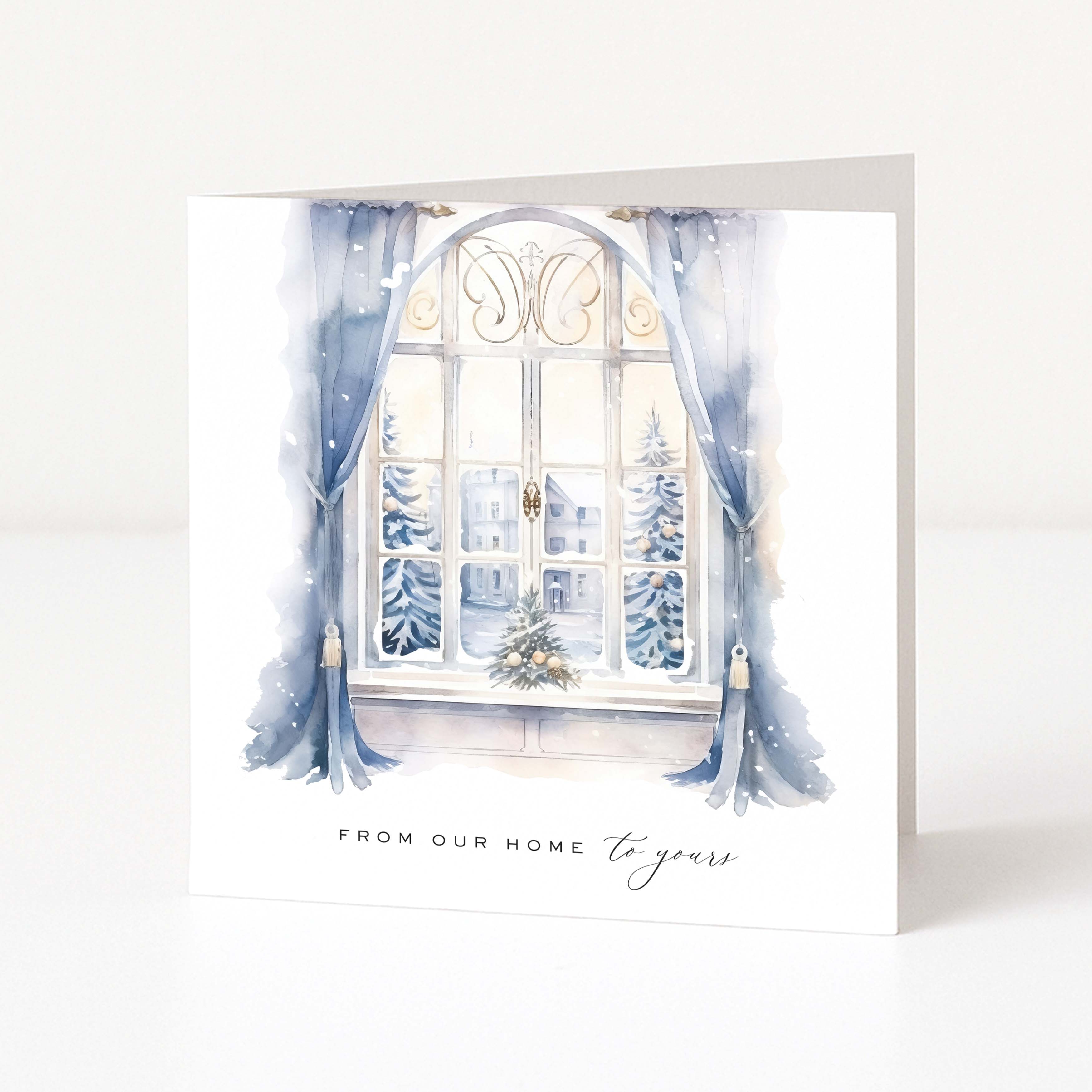 A square greeting card with a watercolor illustration of a snowy window scene, featuring blue curtains, snow-covered trees, and the text 'FROM OUR HOME to yours' at the bottom, shown standing against a white background.