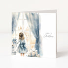 A square Christmas greeting card with a watercolor illustration of a young girl in a blue dress standing by a window with blue curtains, looking out at buildings, with wrapped gifts on the floor. The right side of the card has 'Happy Christmas' printed in black script.