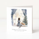 A square white greeting card standing upright on a light gray surface. The front features a watercolor illustration of a girl sitting by a window next to a decorated Christmas tree, with the text 'Wishing you a Merry Christmas AND A HAPPY NEW YEAR' below.