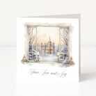 A white greeting card standing upright on a white surface, featuring a watercolor winter scene printed on the front: an open window view of a castle at dusk, snow-covered trees, and the words 'Peace, Love, and Joy' in black script below.