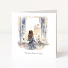 A square greeting card with a watercolor illustration of a girl in a blue dress looking out of a window onto a snowy city street, with the text 'Warmest Winter Wishes' printed below. The card is shown standing upright on a white surface against a light gray background.