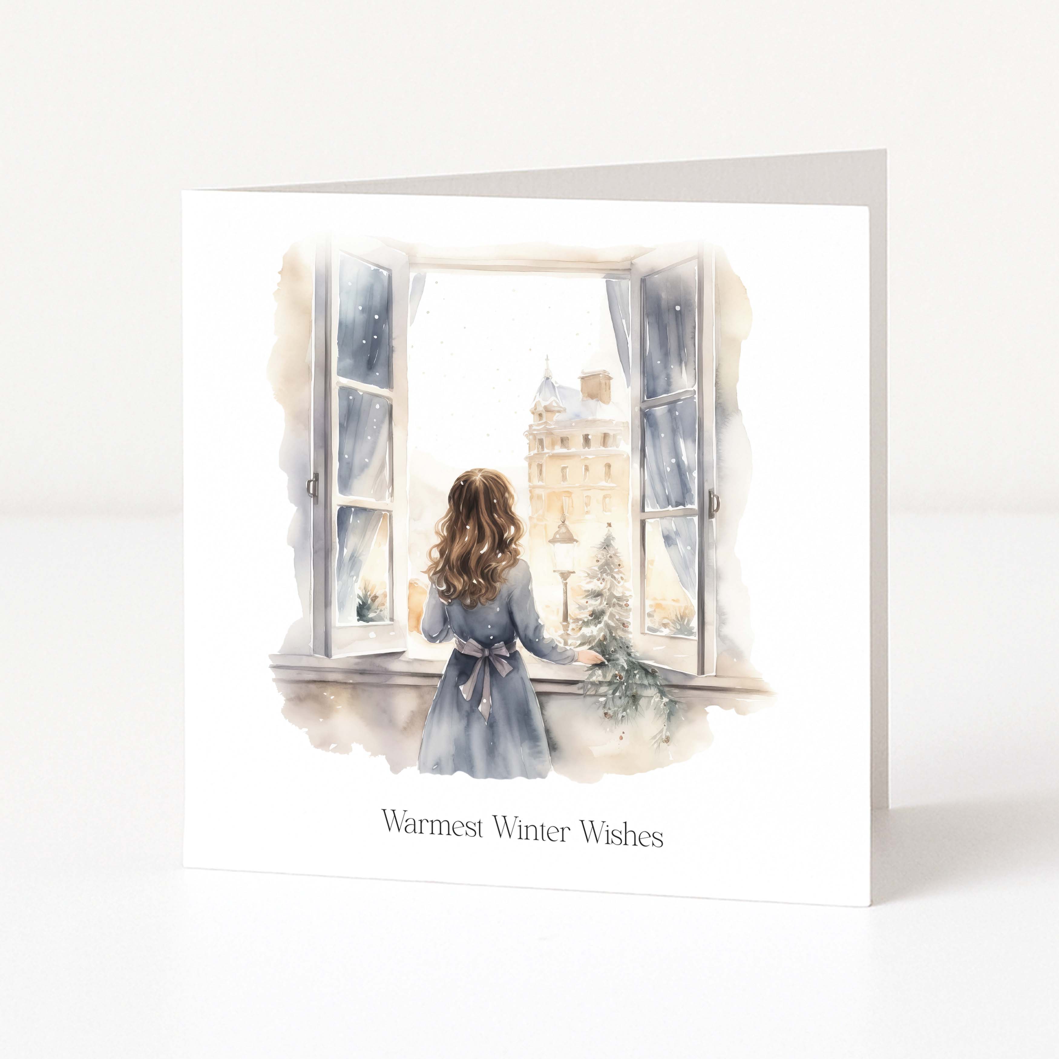 A square greeting card with a watercolor illustration of a girl in a blue dress looking out of a window onto a snowy city street, with the text 'Warmest Winter Wishes' printed below. The card is shown standing upright on a white surface against a light gray background.