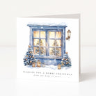 A square Christmas greeting card standing upright on a white surface. The card features an illustrated snowy window with warm yellow light inside, decorated blue Christmas trees, lanterns, and the text 'WISHING YOU A MERRY CHRISTMAS from our home to yours'.