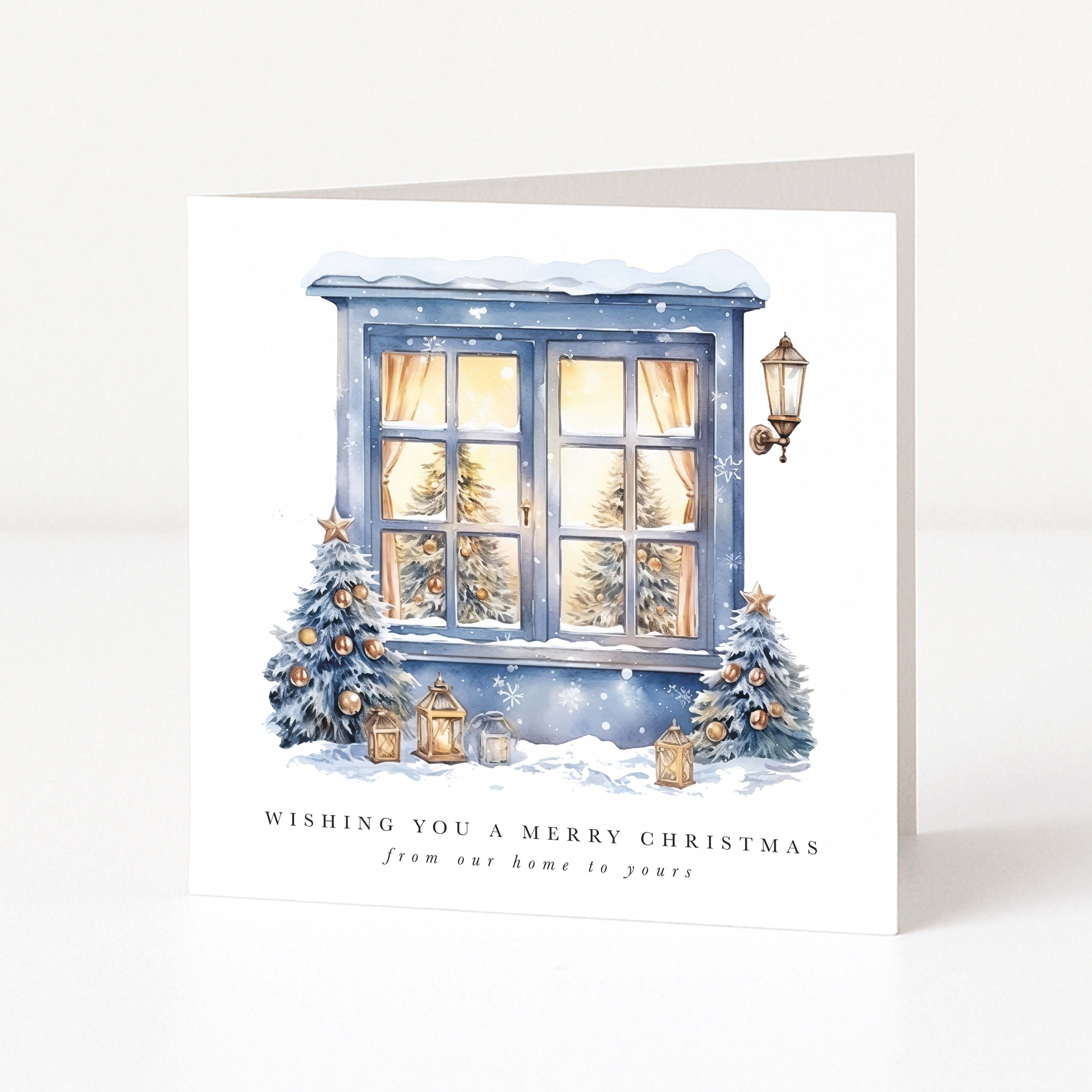 A square Christmas greeting card standing upright on a white surface. The card features an illustrated snowy window with warm yellow light inside, decorated blue Christmas trees, lanterns, and the text 'WISHING YOU A MERRY CHRISTMAS from our home to yours'.