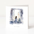 A white greeting card standing upright on a white surface, featuring a watercolor illustration of a woman in a blue dress sitting by an open window and looking out at a snowy city scene. The card is labeled 'let it snow' at the bottom.