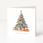 A square white Christmas card standing upright on a white surface, featuring a watercolour-style Christmas tree with ornaments and wrapped gifts beneath it, plus the text 'Jingle all the way' near the bottom.
