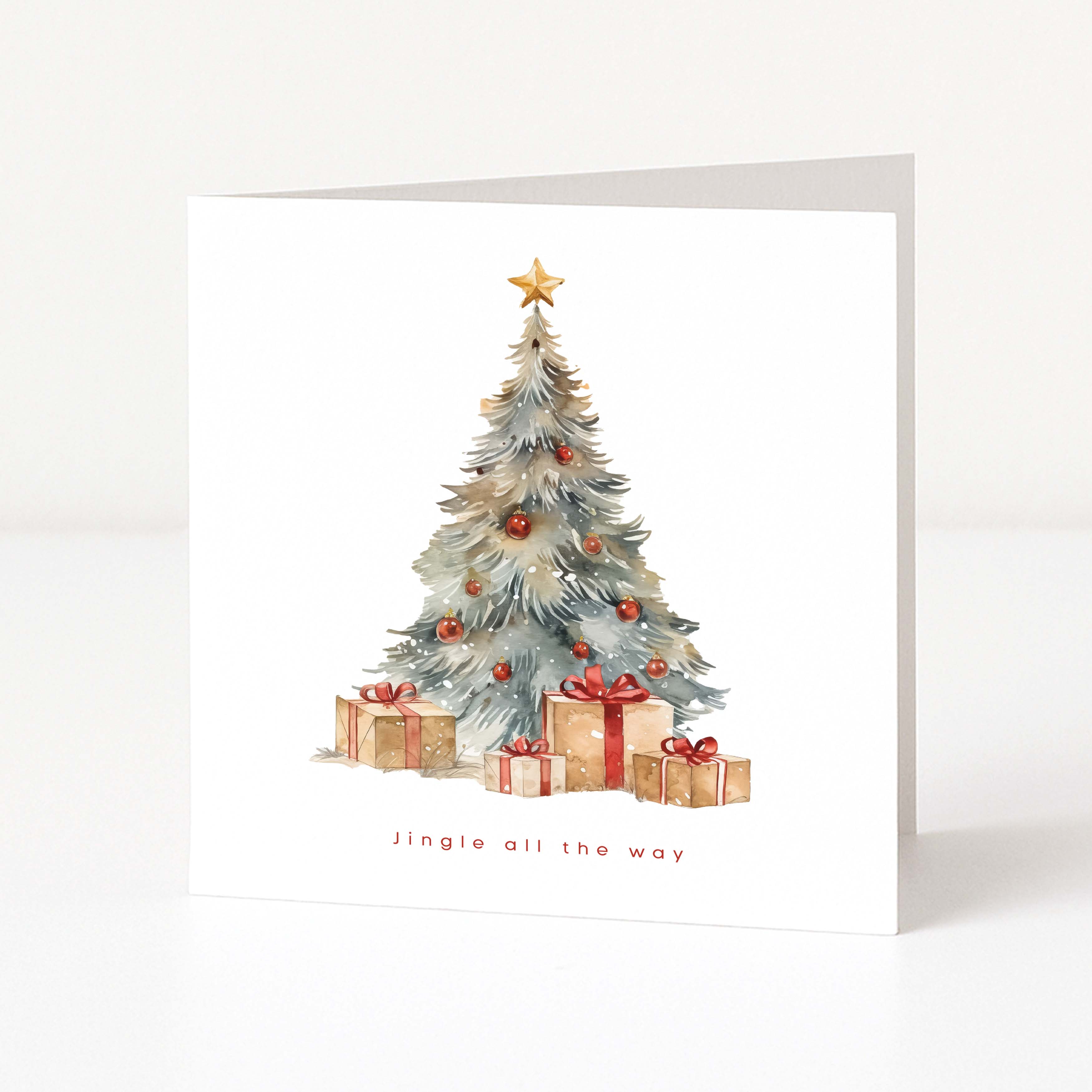 A square white Christmas card standing upright on a white surface, featuring a watercolour-style Christmas tree with ornaments and wrapped gifts beneath it, plus the text 'Jingle all the way' near the bottom.