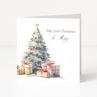 A white Christmas greeting card standing upright, featuring a watercolor illustration of a decorated tree with wrapped presents and the text 'May your Christmas be Merry'.