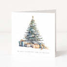 A square white Christmas greeting card standing upright on a light grey surface, featuring a watercolour illustration of a decorated Christmas tree with wrapped presents underneath and the text 'the most wonderful time of the year' near the bottom.