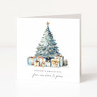 A square white Christmas greeting card standing upright on a white surface, featuring a watercolor illustration of a decorated Christmas tree with wrapped presents and the text 'SEASON'S GREETINGS' and 'from our home to yours'.