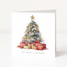 A square white Christmas greeting card standing upright on a light surface. The front features a watercolor-style decorated tree with a gold star, red and gold ornaments, wrapped presents, and the text 'EAT, DRINK and BE MERRY'.