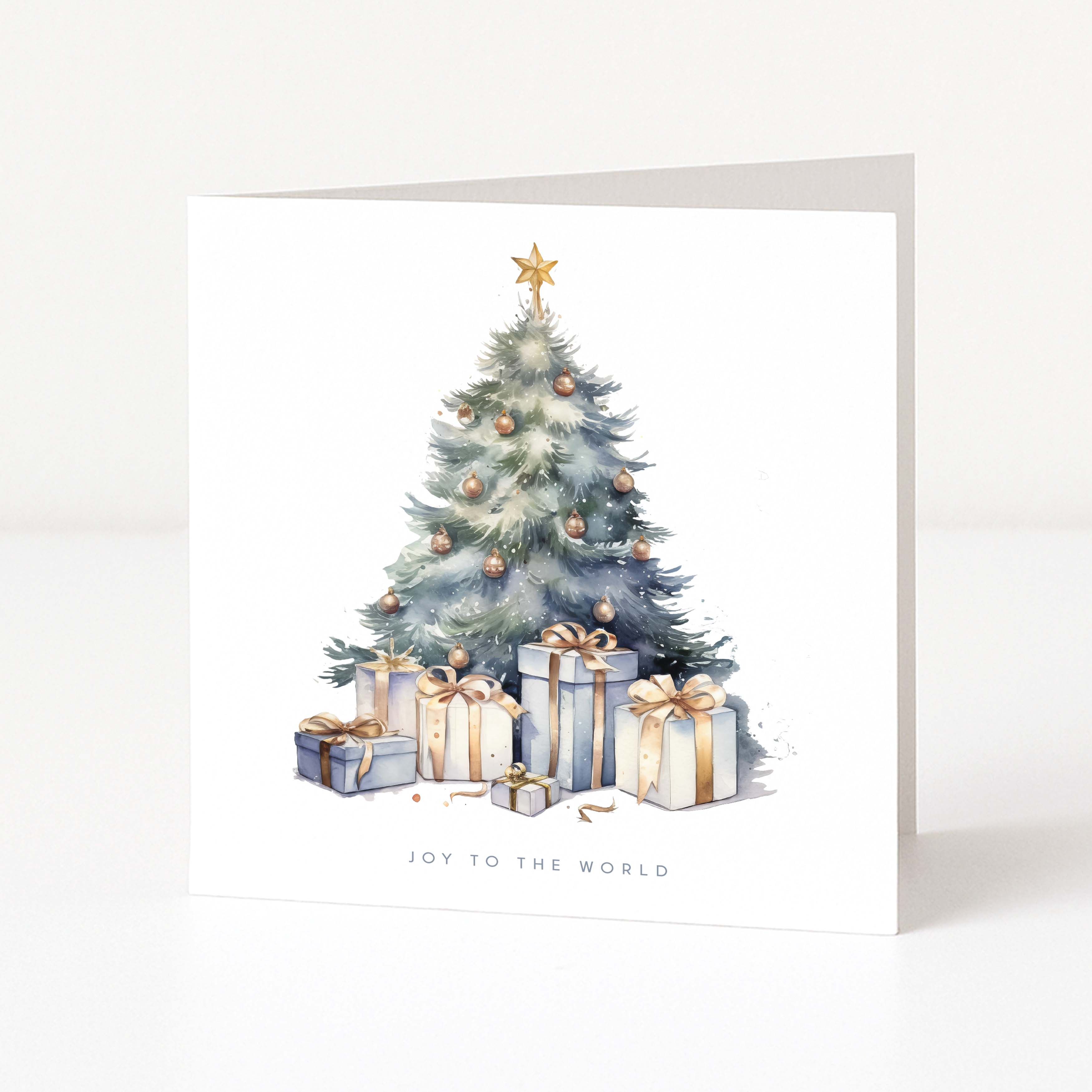 A square white Christmas greeting card standing upright on a light surface, featuring a watercolour-style Christmas tree with gold ornaments and several wrapped presents beneath it. The card reads 'JOY TO THE WORLD' at the bottom.